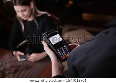 The waitress's hands hold the terminal for payment. Close-up photo. The view from the top. The guest sits at the table with the phone out of focus. The concept of service in the restaurant.