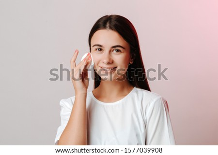 Happy teen girl caring about her healthy face skin with cotton disk, lookin at the camera and smiling on grey background.
