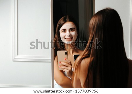 Girl with face clay mask taking a selfie in a mirror on a smartphone. Self care and skin treatment.