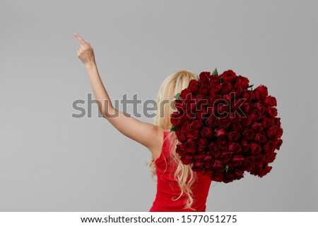 Happy young blond woman holding a large bouquet of red roses as a gift for March 8 or Valentine. Pointing left to empty space for text. Neutral gray studio background