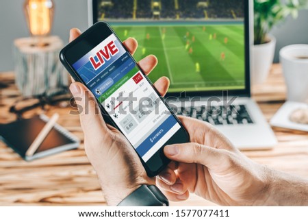 Close up cropped shot of male hands betting online using gambling mobile application on his phone. Man watching football match online broadcast on his laptop waiting for winning results.
