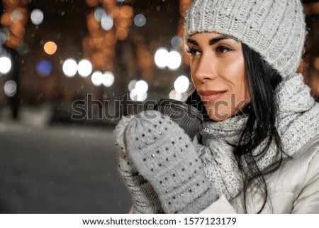 Beautiful girl wearing in wool clothing holding coffee cup on background of night street. Lady looking away. Stylish knitted hat, gloves and scarf warming young woman in winter.