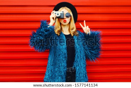 Portrait stylish woman with retro camera taking picture blowing red lips sending sweet air kiss showing peace gesture wearing blue faux fur coat over red wall background