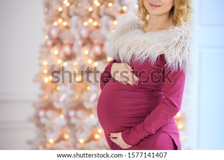 pregnant blonde woman in purple dress holds her arms around the round stomach on christmas tree background in white room, holiday concept, closeup. Expecting baby
