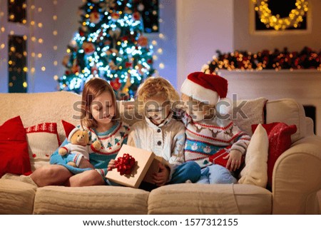 Children at Christmas tree and fireplace on Xmas eve. Family with kids celebrating Christmas at home. Boy and girl in knitted sweaters on white couch opening presents. Holiday gifts for kid.