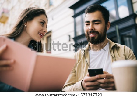 Low angle of young female demonstrating notepad with data to bearded ethnic colleague with smartphone while doing remote job on city street