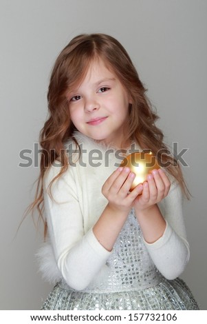 Caucasian beautiful little girl in a shiny dress with cute yellow hair holding a Christmas ball on a gray background/Christmas and New Year decoration