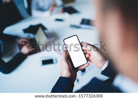 From above crop businessman in suit using smartphone with blank screen to search information in internet during meeting in office