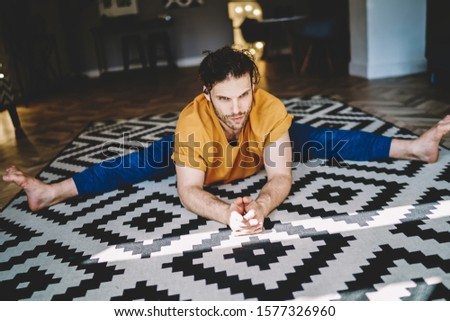 From above of young athletic strong male in orange t shirt and blue pants stretching legs and arms lying on white and black carpet in room 
