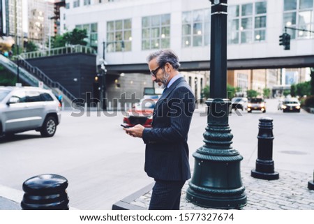 Side view of handsome concentrated gray haired businessman in glasses and black costume tapping on smartphone in New York on blurred background
