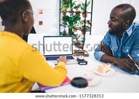 Side view of focused concentrated African American colleagues sitting in coffee shop and working with laptop with white blank screen while discussing business issues