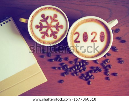 White coffee cup with number 2020 over frothy surface on brown wooden background with another one with snowflake sign, coffee beans, blank calendar. Holidays food art creative theme for New Year 2020.