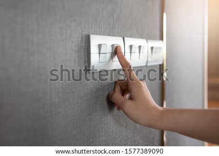 Asian female right hand is turning on or off on grey light switch over gray textile texture wall at the house or hotel. Copy space. Royalty-Free Stock Photo #1577389090