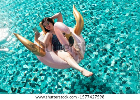 Beautiful glamorous young brunette woman taking a selfie on a pink and gold flamingo / swan pool float in the swimming pool on a sunny day