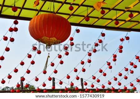 One big red Chinese lantern with background of aligned of small lanterns hanging. Sky is blue.