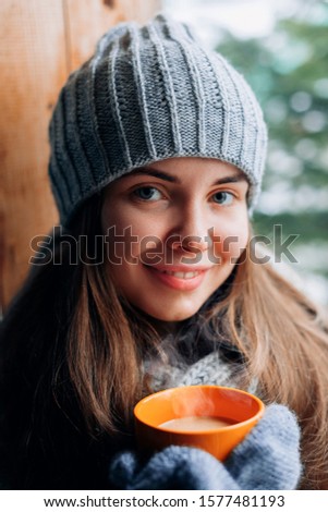 Beautiful woman holding and drinking a cup of coffee or cocoa in gloves sitting home by the window. Blurred winter snow tree background. Morning, coziness, winter and people concept 