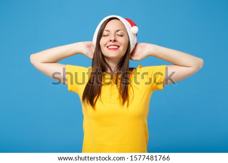 Calmed young brunette woman Santa girl in yellow t-shirt Christmas hat posing isolated on blue background. New Year 2020 celebration holiday concept. Mock up copy space. Sleep with hands behind head