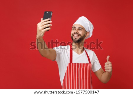 Smiling young chef cook or baker man in striped apron toque chefs hat isolated on red wall background. Cooking food concept. Mock up copy space. Doing selfie shot on mobile phone, showing thumb up