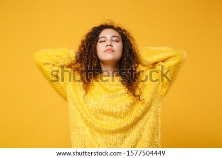 Relaxed young african american girl in fur sweater posing isolated on yellow orange wall background, studio portrait. People lifestyle concept. Mock up copy space. Sleeping with hands behind head