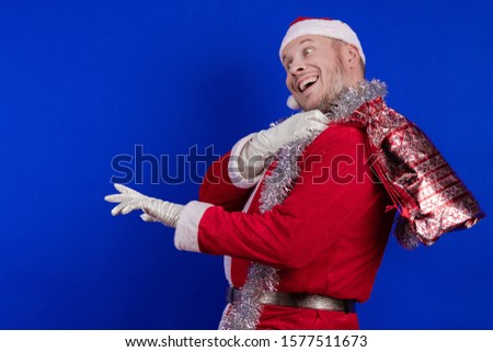 Emotional male actor in a costume of Santa Claus holds a bag with gifts in his hands and poses on a blue background