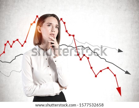 Worried young businesswoman standing near concrete wall with falling graphs. Concept of financial crisis and loss.