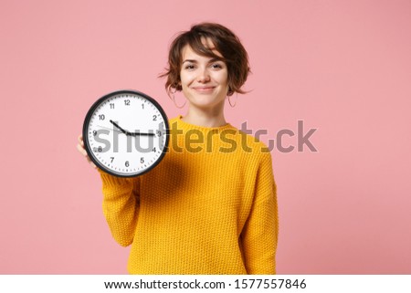 Smiling young brunette woman girl in yellow sweater posing isolated on pastel pink wall background, studio portrait. People sincere emotions lifestyle concept. Mock up copy space. Hold in hand clock
