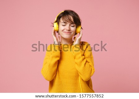 Relaxed young brunette woman girl in yellow sweater posing isolated on pastel pink background in studio. People lifestyle concept. Mock up copy space. Listen music with headphones keeping eyes closed