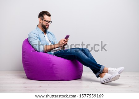 Full length profile photo of positive guy sitting comfy violet armchair holding telephone chatting friends wear specs casual denim outfit isolated grey color background Royalty-Free Stock Photo #1577559169