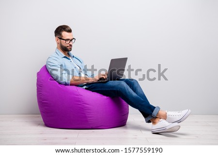 Full length profile photo of attractive guy holding notebook browsing chatting colleagues sitting comfy soft armchair wear specs casual denim outfit isolated grey color background
