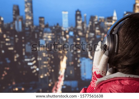 Young woman in a winter jacket and Christmas knitted gloves listening music in the headphones on roof of night New York. Aerial and panorama view of skyscrapers
