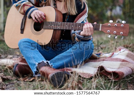 Young blond woman, wearing colorful cardigan and blue jeans, holding acoustic guitar, sitting on blanket in countryside in autumn. Close-up picture of guitar in hands of Hippie musician
