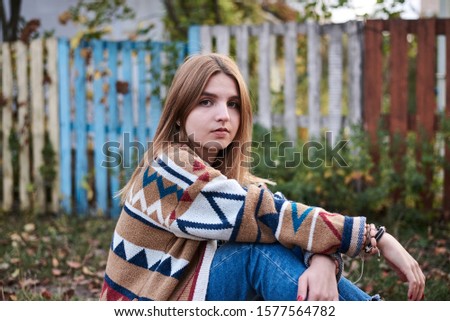 Young blond woman, wearing colorful cardigan and blue jeans, sitting on blanket by colorful old wooden fence in countryside. Hippie musician posing for picture in the park in autumn fall.