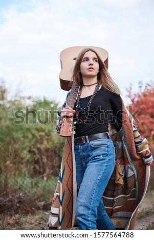 Young blond woman, wearing colorful cardigan and blue jeans, holding acoustic guitar on her shoulder, walking in park woods forest in autumn. Three-quarter portrait of hippie musician with guitar.
