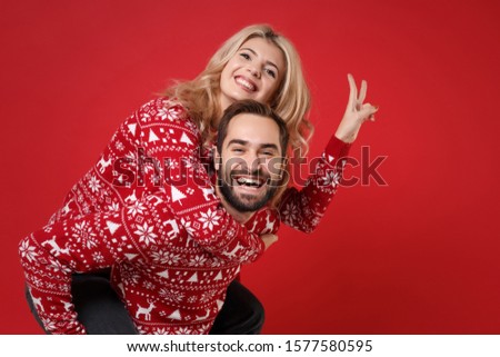 Smiling young couple guy girl in Christmas sweaters posing isolated on red background. Happy New Year 2020 celebration party concept. Mock up copy space. Giving piggyback ride to joyful sit on back