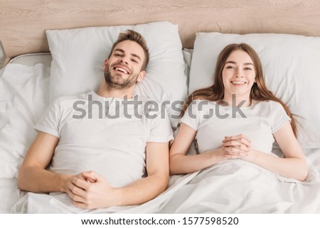 top view of cheerful husband and wife lying in bed and smiling at camera