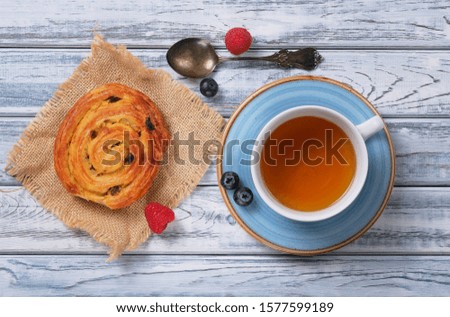 Sweet puff bun with cream and raisins in the shape of a snail on burlap. Tasty delicious dessert, silver old teaspoon, cup of tea, berries of fresh juicy raspberries and blueberries. French breakfast.