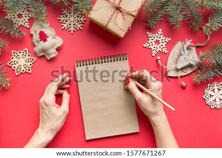 Zero waste Christmas concept with wooden natural decor on red. Woman writing invitation. Goals, checklist, plans, wish list, to do list for New Year. Top view. Flat lay.