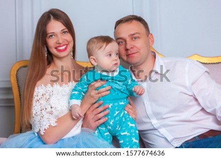 Young family are sitting by the sofa in a cozy room. The son sitting between the parents looks in the frame and smiles