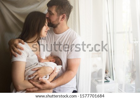 Father and mother with a newborn son standing by the window. New young multi-generation small family. Breast-feeding home concept Royalty-Free Stock Photo #1577701684