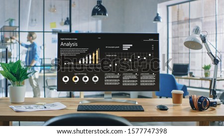 Shot of a Desktop Computer in the Creative Modern Office. Monitor Screen Shows Company Growth Data with Graphs, Charts, Software UI. In the Background Young Professional Looking for a Book. Royalty-Free Stock Photo #1577747398