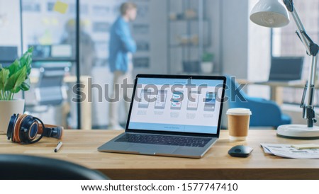 Shot of Laptop Standing at Desk in the Creative Office. Notebook Screen Shows Mobile Phone Application Design, Software UI Development. In Background Young Professional Stans in Creative Modern Office