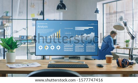 Shot of a Desktop Computer in the Creative Modern Office. Monitor Screen Shows Company Growth Data with Graphs, Charts, Software UI. In the Background Young Professional Using Smartphone. Royalty-Free Stock Photo #1577747416