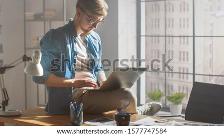 Professional Creative Designer Sits on His Desk Holds Laptop on the Knees and Working on the Project Early Morning. Creative Designe and Gaming Content Development Studio