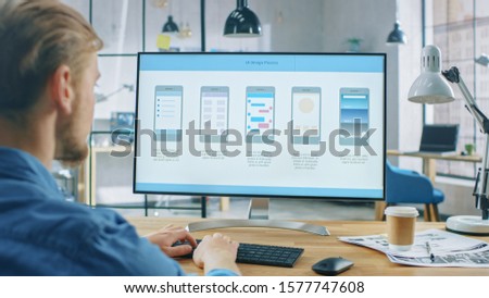 Young Professional Works on Desktop Computer at His Desk in the Creative Office. Monitor Screen Shows Mobile Phone Application Design, Software UI Development. In the Background Creative Modern Office