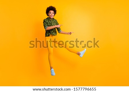 Full length photo of funny dark skin curly lady jumping high indicating fingers empty space low prices banner wear green dotted shirt pants isolated yellow color background