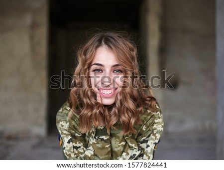Young curly blond military woman, wearing ukrainian military uniform, posing for picture. Three-quarter portrait of soldier in front of ruined abandoned building, construction site.