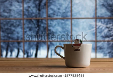 Christmas and New Year Simple Home Decor: Mug and Santa Claus with Christmas Tree on the Wooden Table against Blurry Window with Blue View of Winter Snowy Trees Outside, - Background, Copy Space.