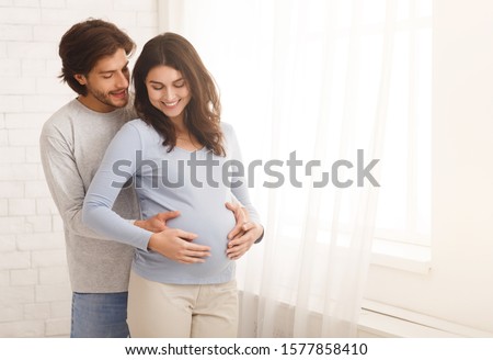 Loving man hugging his pregnant wife from behind standing near window at home, copy space Royalty-Free Stock Photo #1577858410