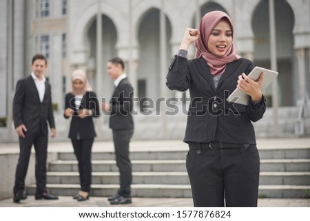 Young Muslim businesswoman reacts while using tablet with team discussing in the background