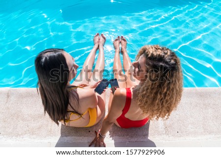 Stock photo of two girls of different ethnic groups sitting at the edge looking at each other and with the mobile in the hand. Summertime